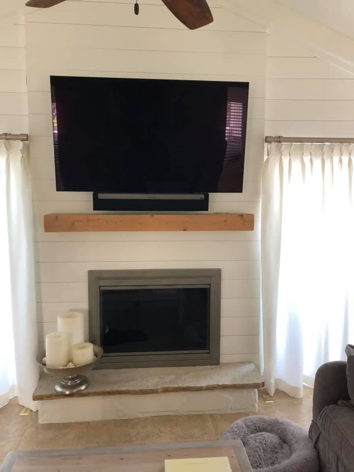Living room with TV above fireplace, white shiplap, light wood mantle, white curtains, and beige floor.