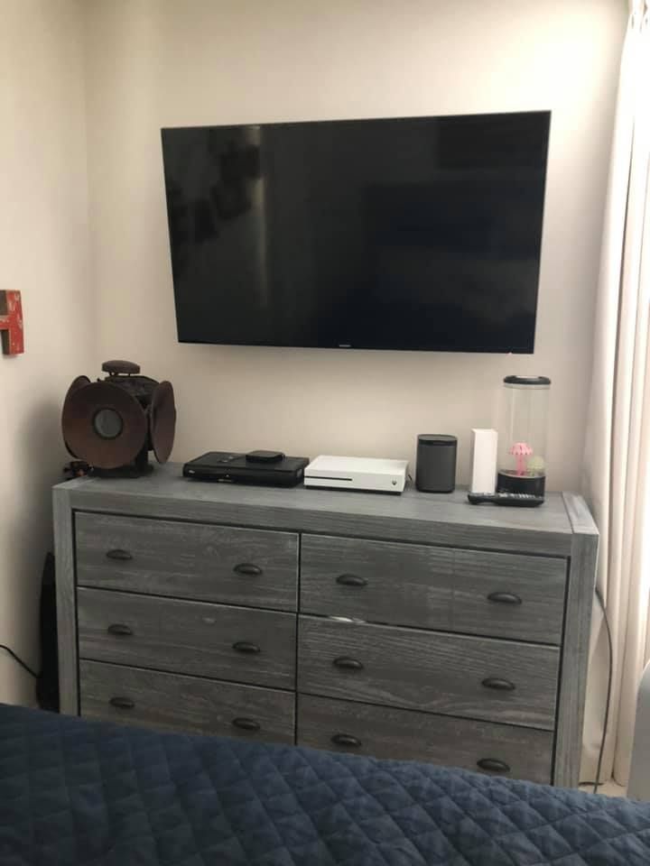 A gray dresser with a large TV mounted above it, with electronics and decorations on top, next to a window.