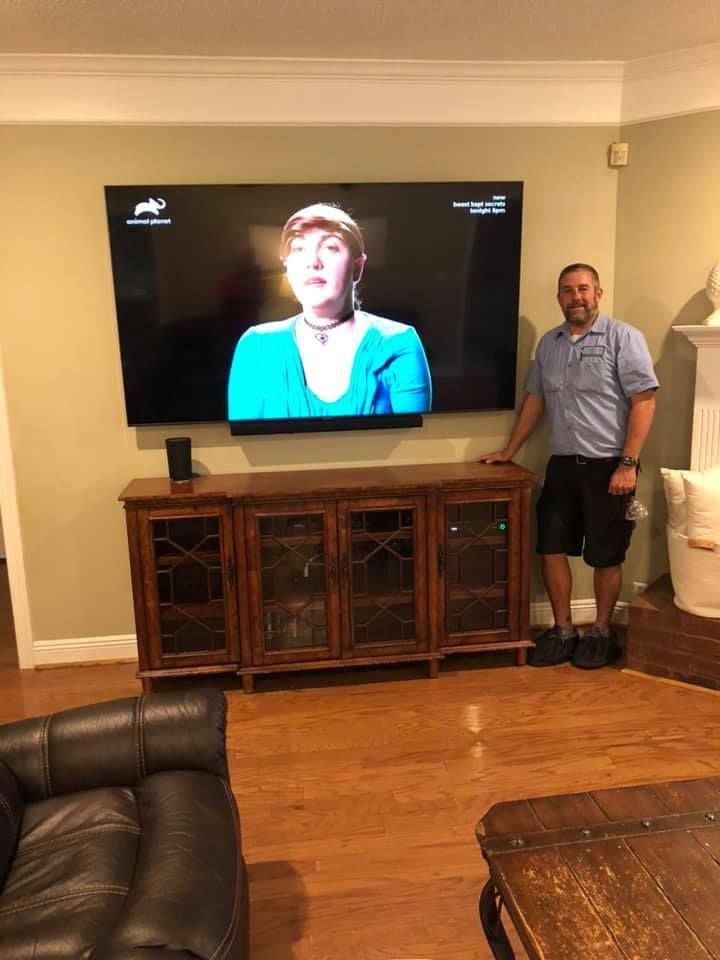 A man stands near a large, mounted TV over a wooden cabinet, set in a living room with hardwood floors.