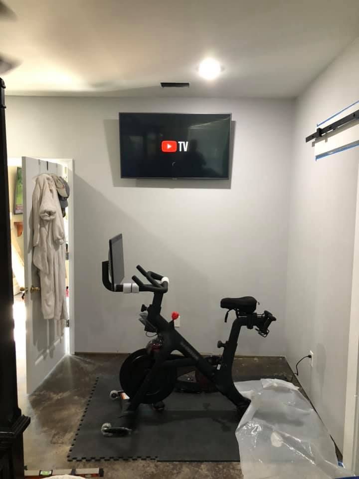 Peloton bike in a home gym with a mounted TV playing YouTube TV. White walls, black mat, and gray flooring.