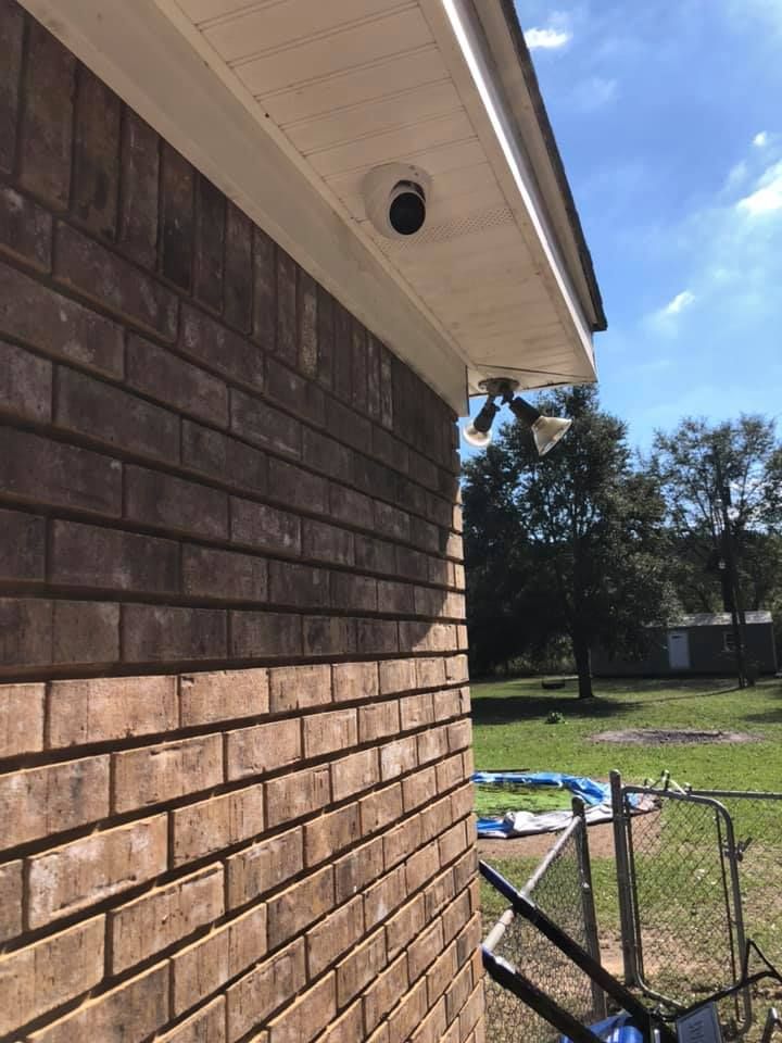 Brick building with security cameras mounted under the white overhang. Green yard and blue sky are visible.