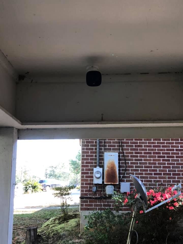A black security camera is mounted on the ceiling of a porch. A brick wall is visible behind it.