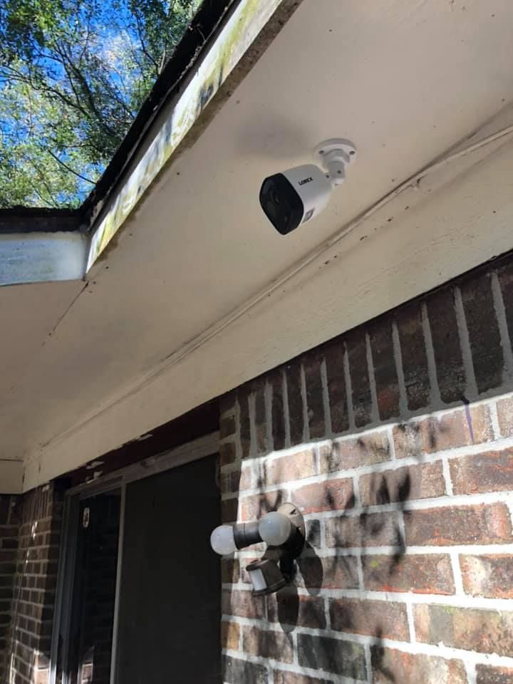 Security camera mounted on a white porch ceiling, above a brick wall with an outdoor light fixture.