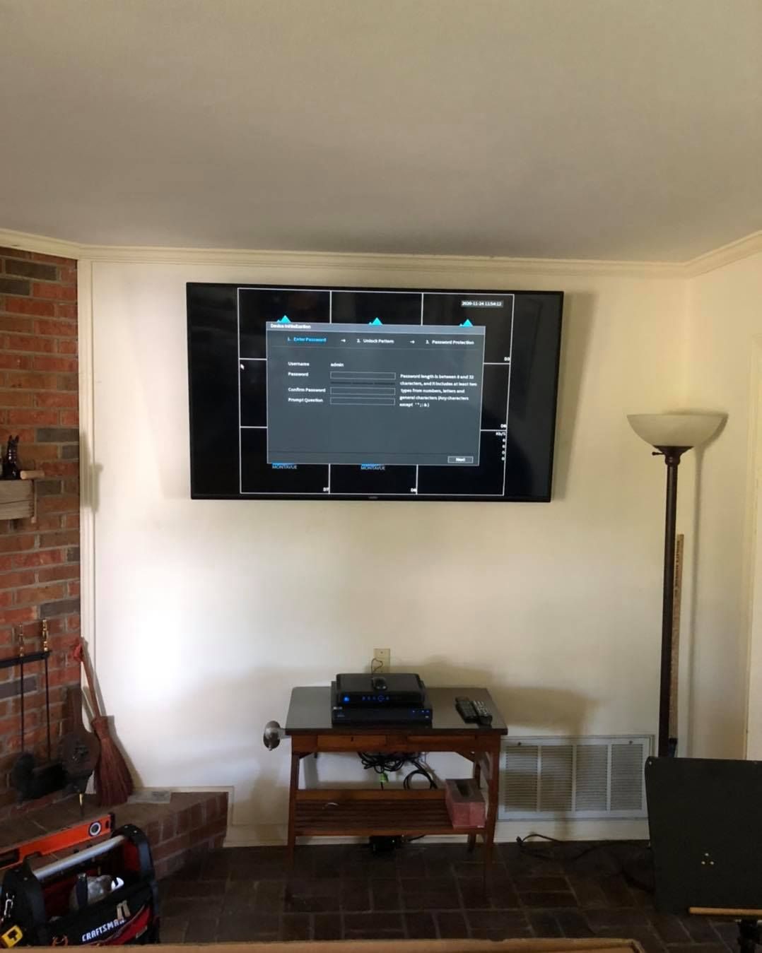 A large TV mounted on a wall with a dark border. A small table holds electronics below. Brick fireplace on the left.