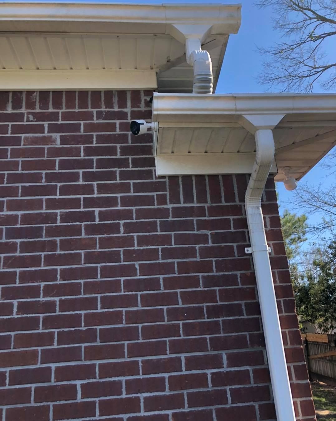 Brick wall with a security camera mounted under white gutters; blue sky.