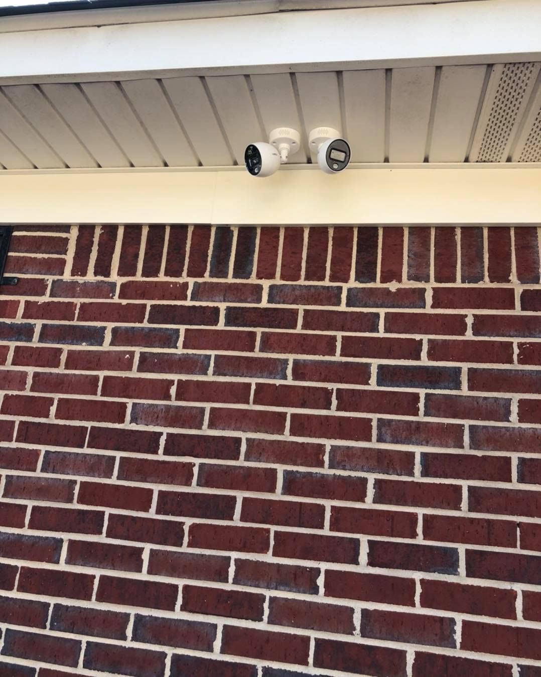 Two white security cameras mounted on the underside of an off-white eave, above a red brick wall.