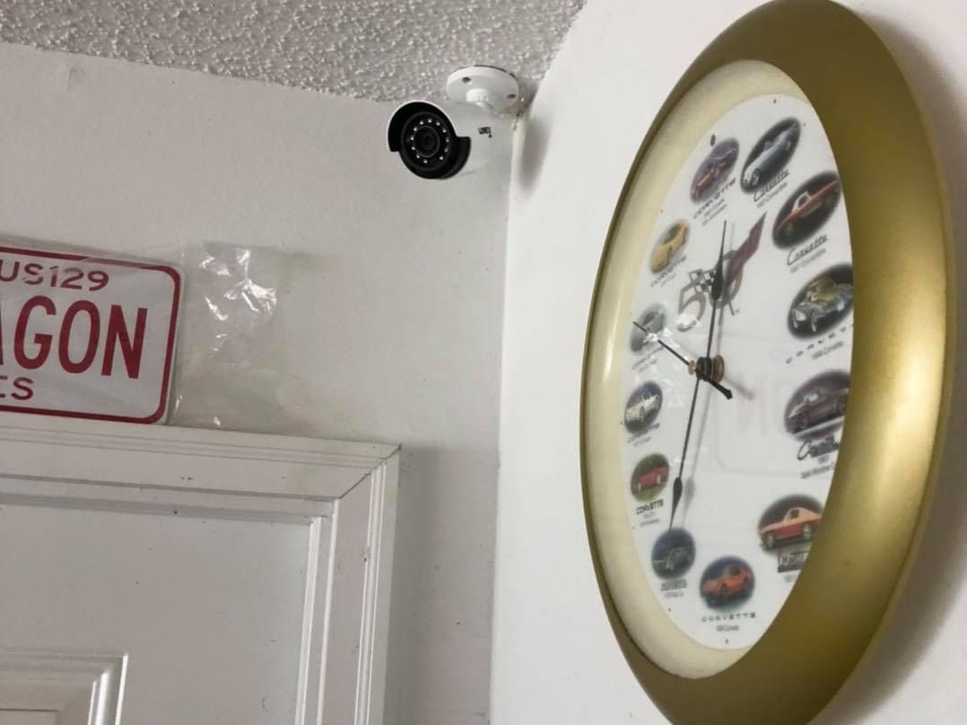 Security camera near a clock and a license plate on a white wall.