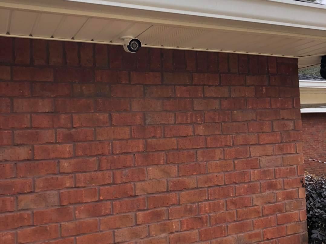 Security camera mounted under white siding on a red brick wall.