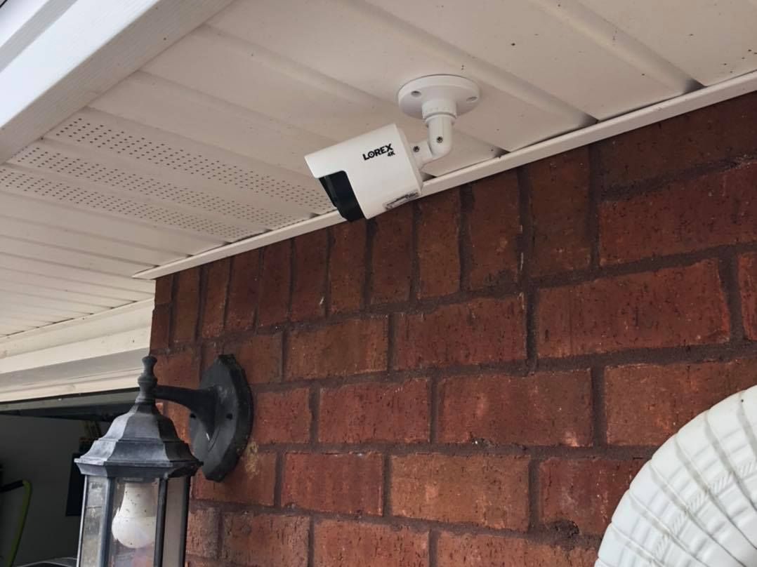 White security camera mounted on a brick wall, next to a black outdoor light fixture, under a white eave.