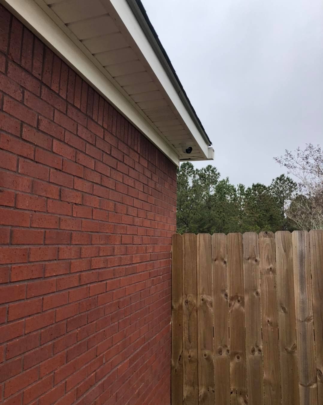 Red brick exterior wall next to a wooden fence; a security camera is mounted under the roof's white overhang.