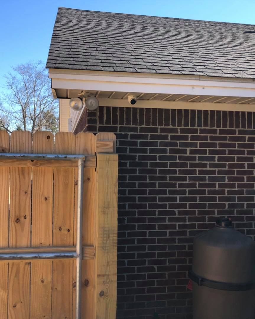 Brick wall with security camera, floodlight, and wooden fence under a shingled roof.