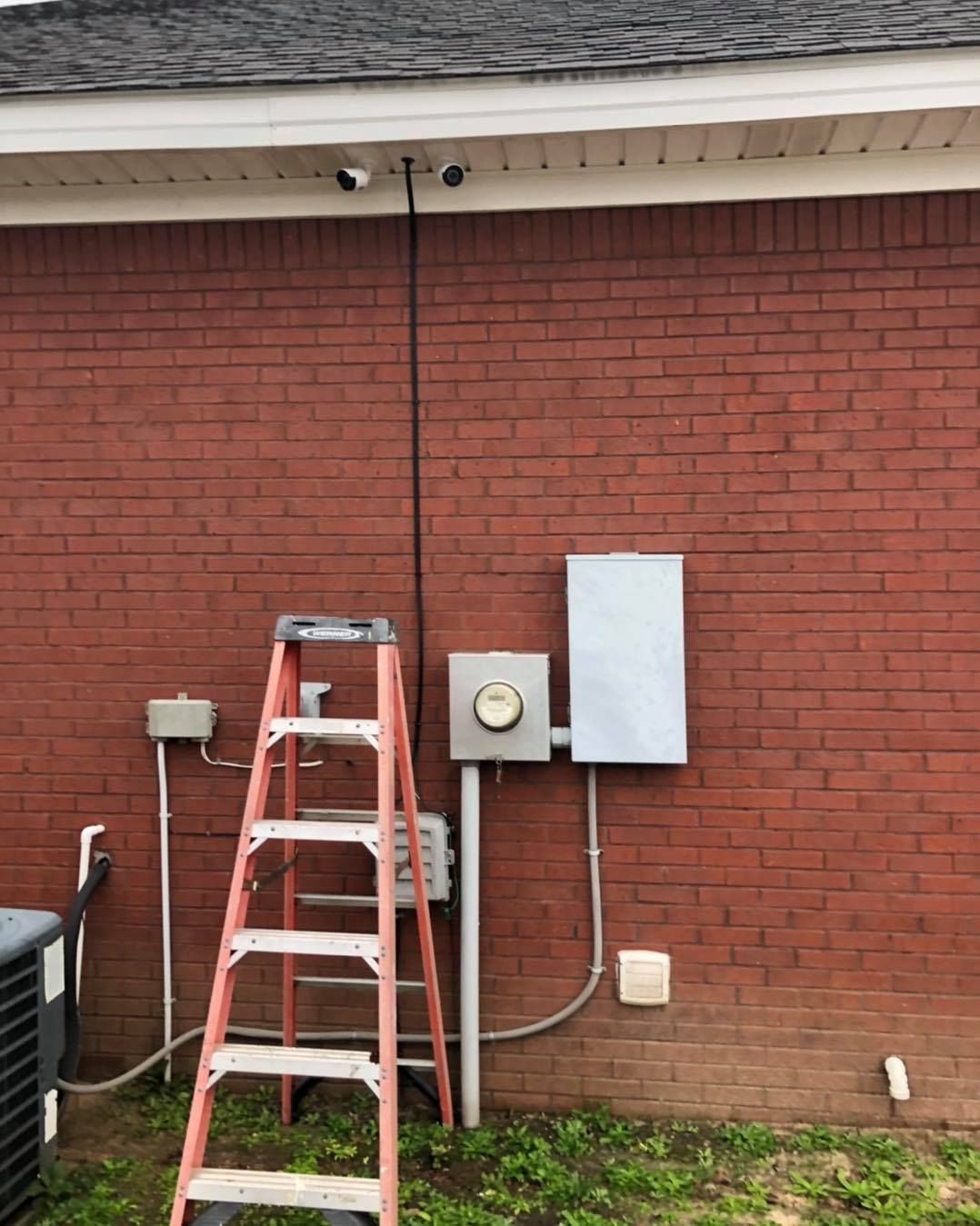 Ladder against red brick wall with security cameras, electrical boxes, and AC unit.