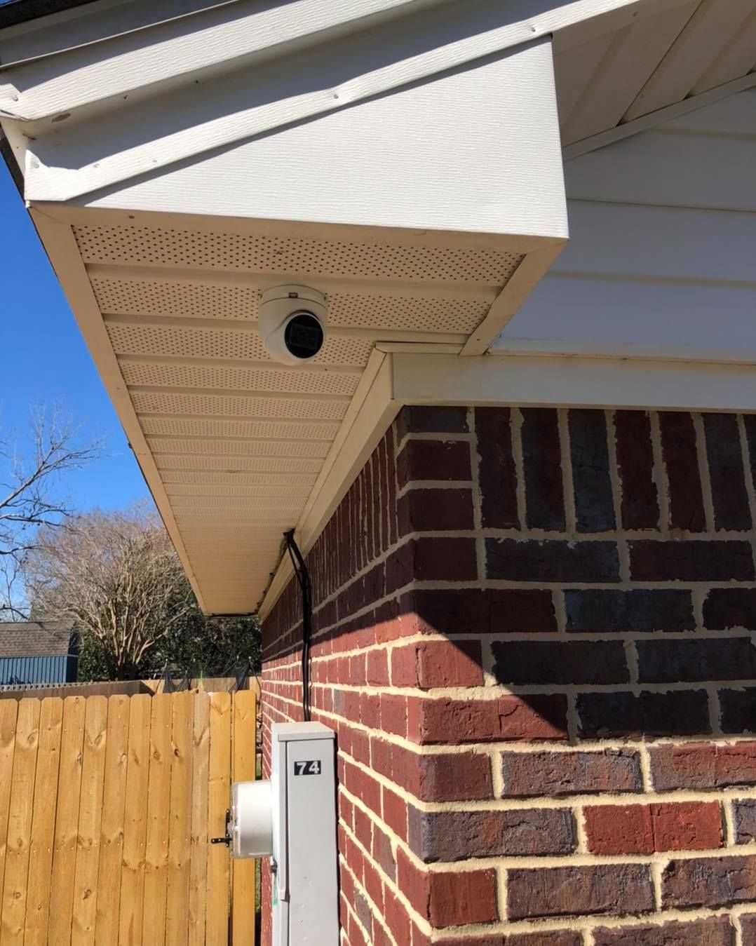 Security camera mounted on the underside of white eaves over a red brick wall.