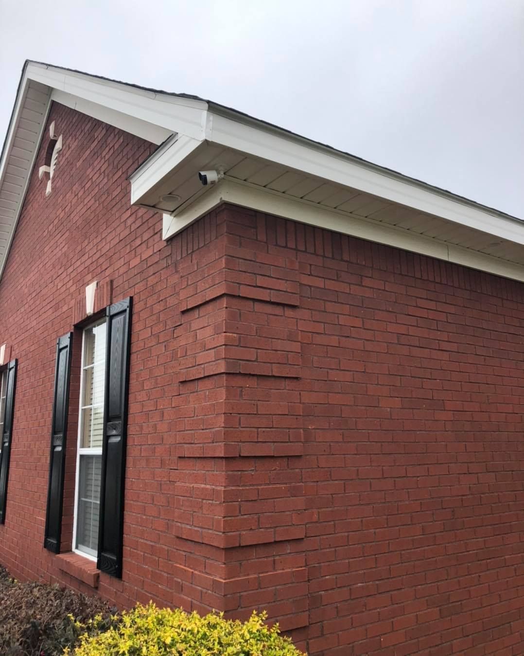 Red brick house corner with white trim, black shutters, and security camera. Overcast day.