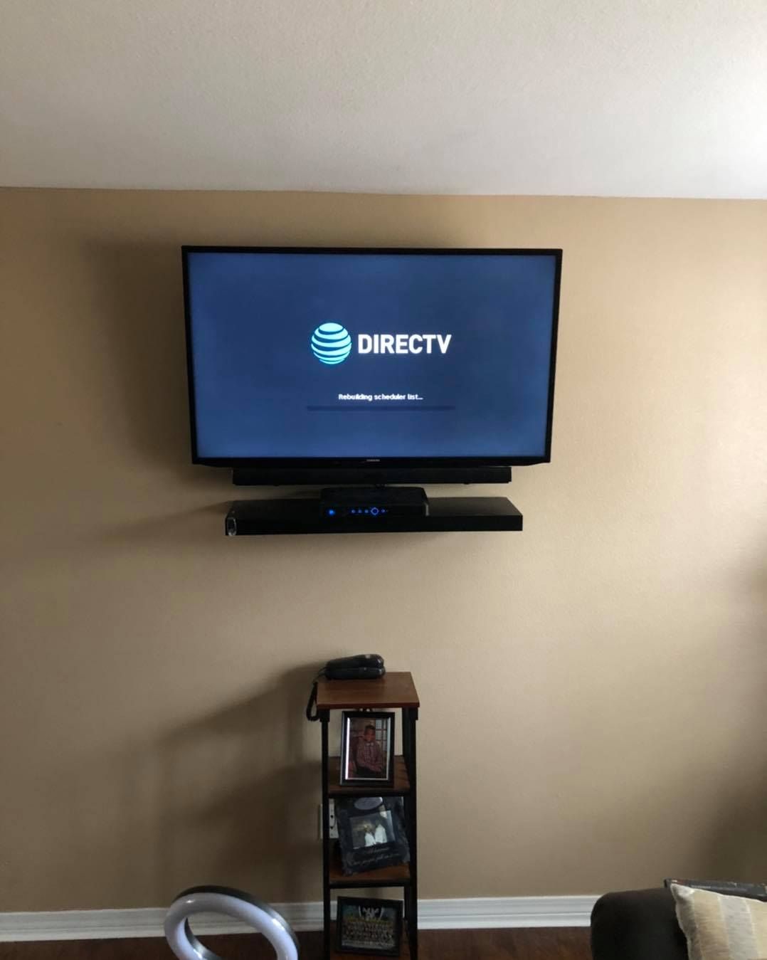 A mounted TV with a soundbar and small shelf on a beige wall above a small wooden shelf.