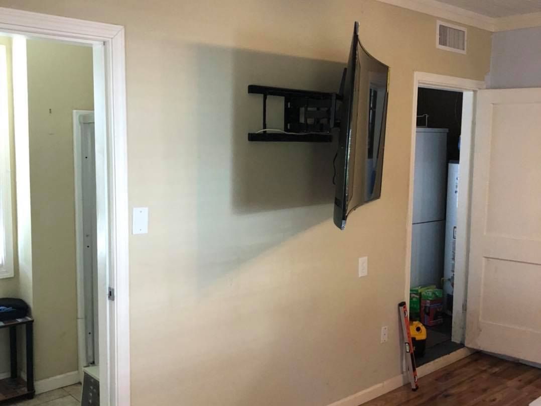 TV mounted on beige wall, doorway to bathroom on left, doorway to room on right with refrigerator visible.