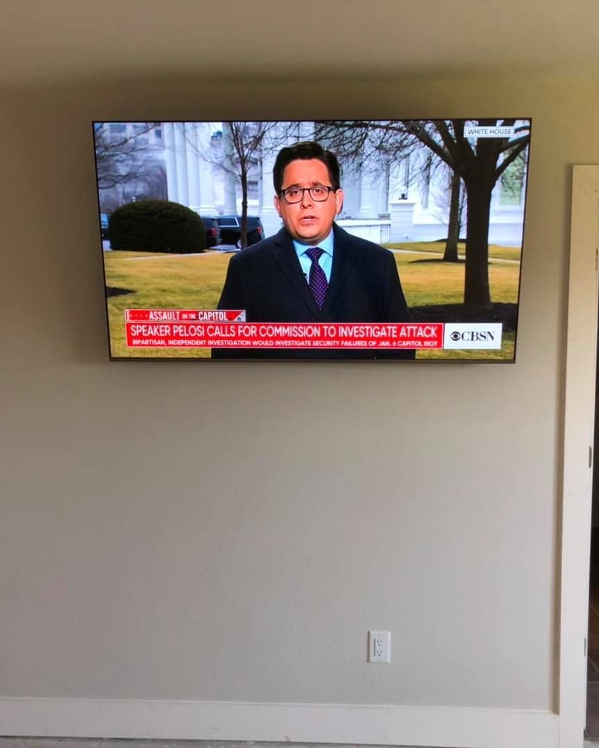 A flat-screen TV mounted on a beige wall displays a news anchor reporting.