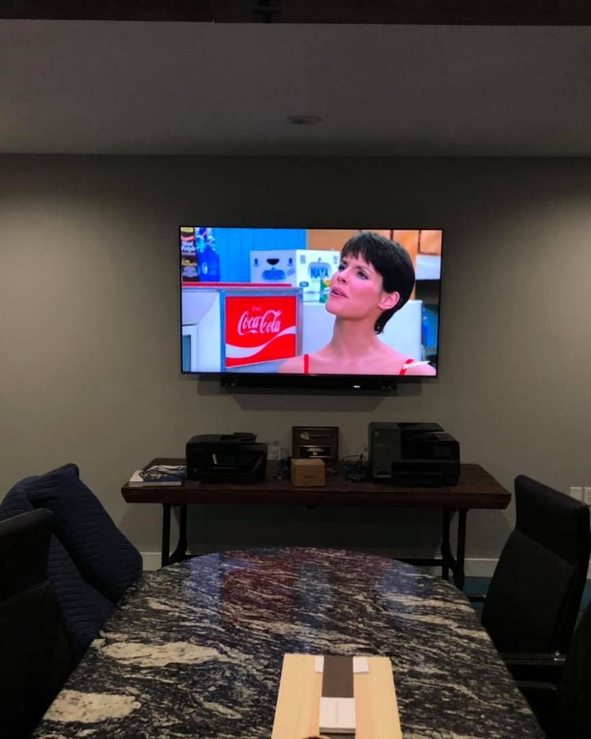 A TV showing a woman in a Coca-Cola ad is mounted on a gray wall. A dark table and a dining table are below.