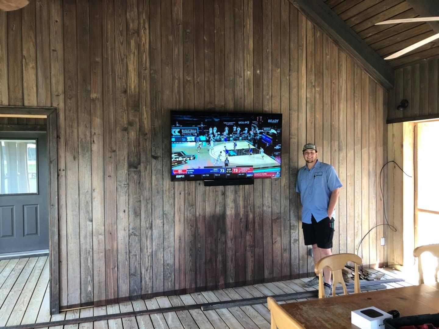 Man standing near a TV mounted on a wood-paneled wall; watching basketball in an outdoor space.