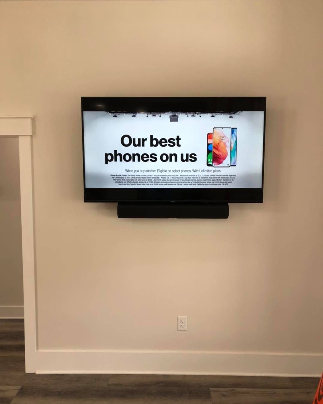 TV on a wall displaying an advertisement for phones. Black soundbar mounted below the TV.