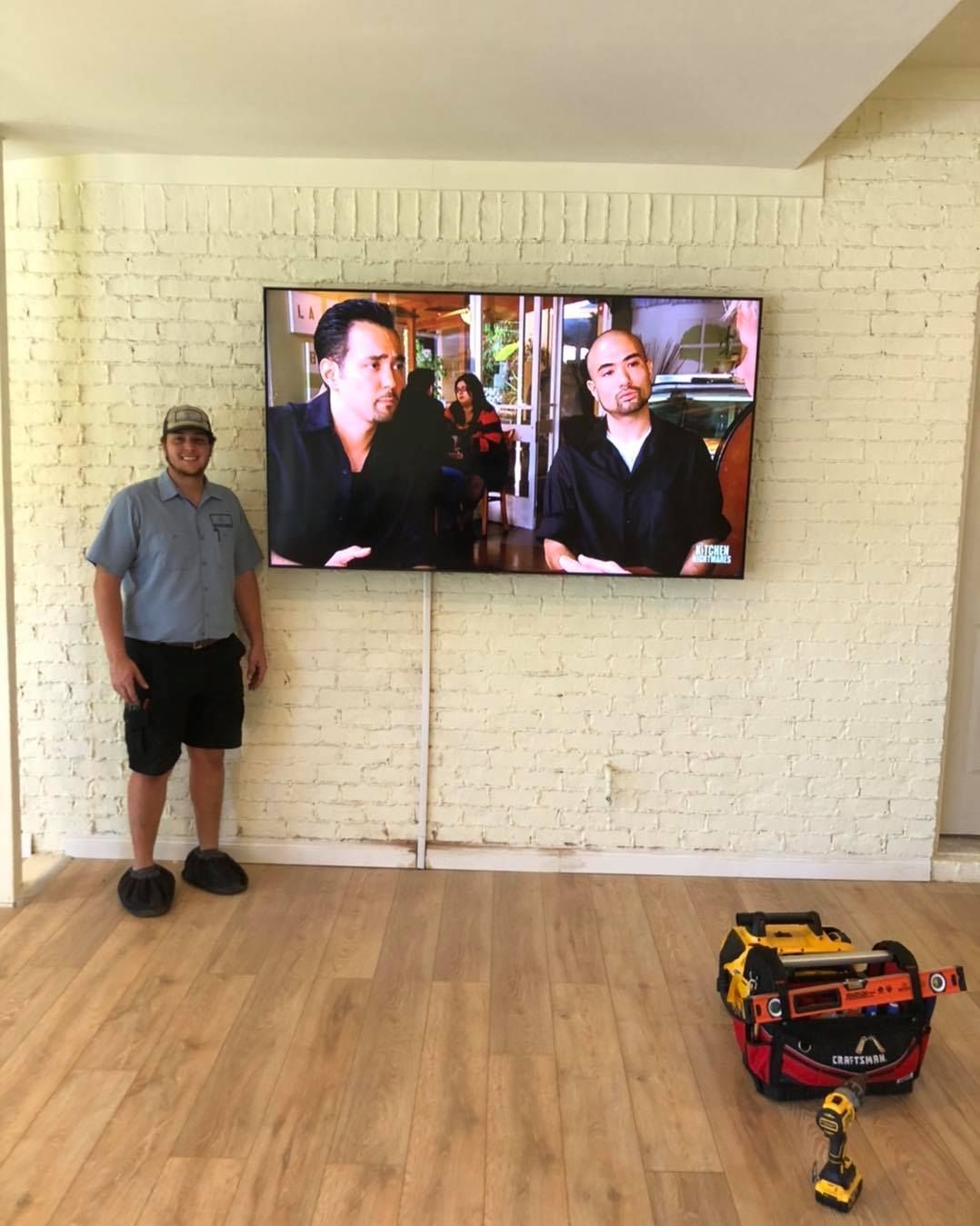 Man stands next to wall-mounted TV on brick, tool bag in front. Wood flooring, bright setting.