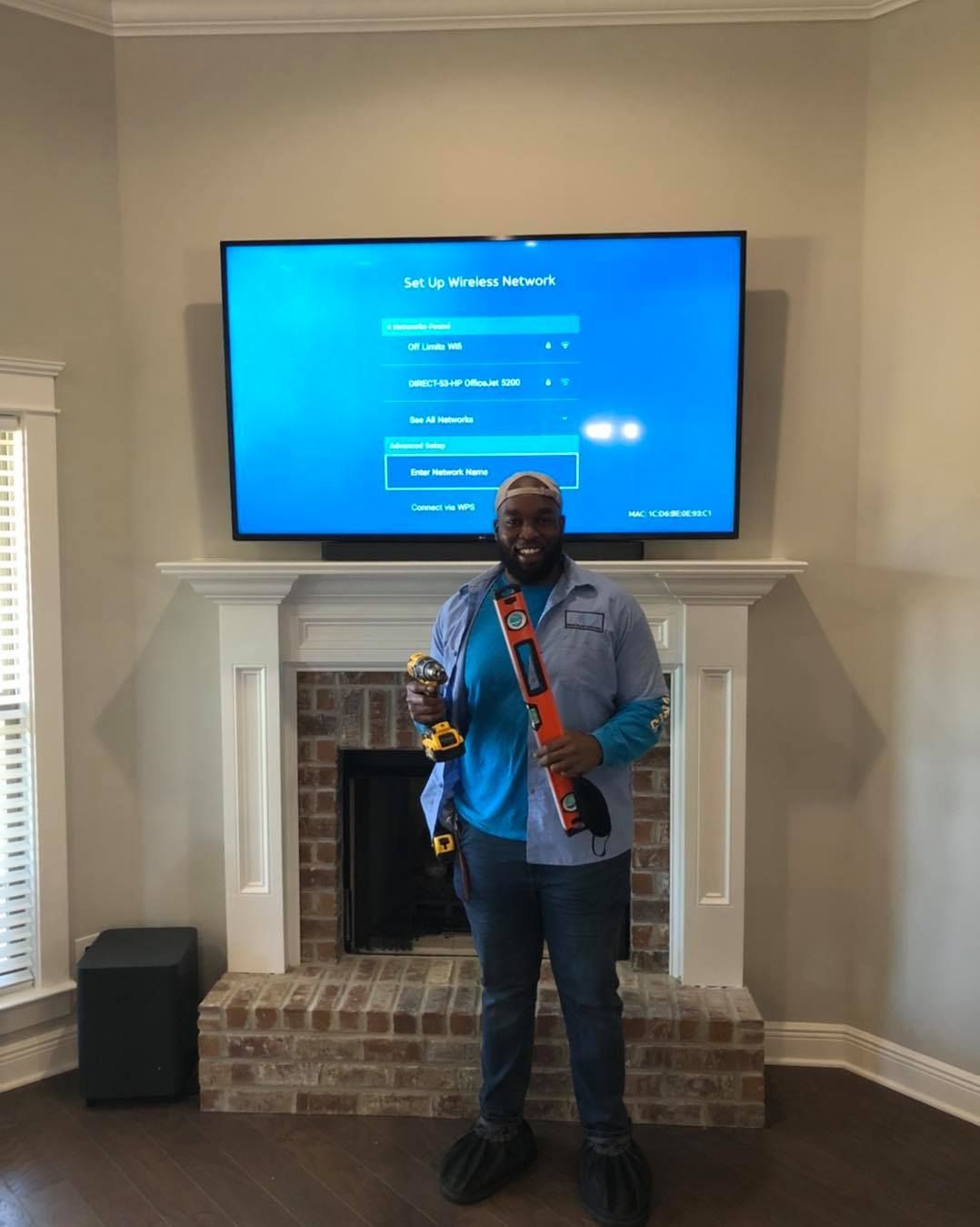 Man smiles, holding tools, in front of a fireplace with a mounted TV. Blue screen, brick, and wood floors.