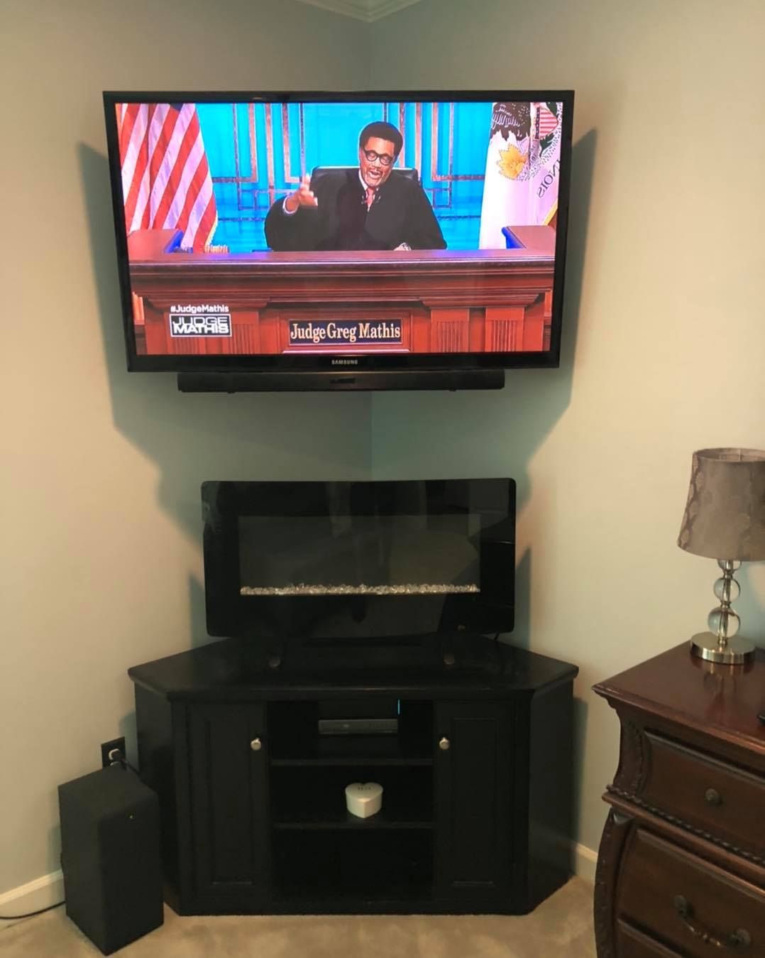 TV with Judge Mathis on mounted above a black entertainment center in a corner, next to a lamp.