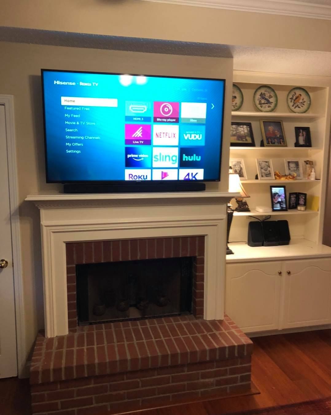 TV over a brick fireplace, showing streaming app menu. White built-in shelving and wooden floor nearby.