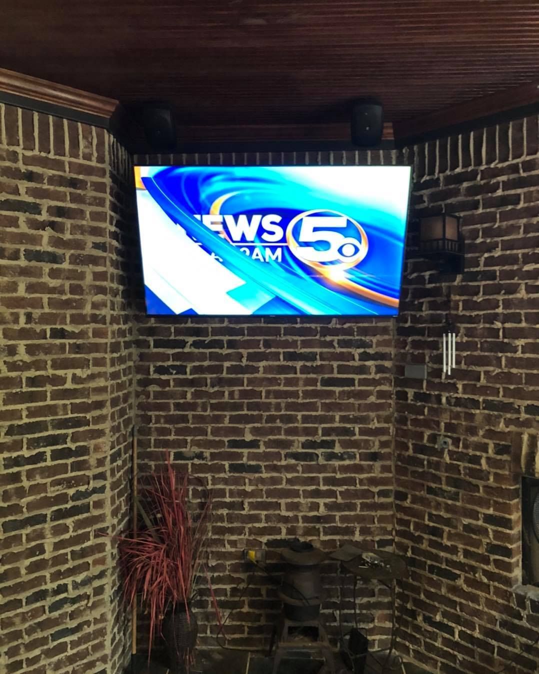TV mounted on a brick wall displaying local news, flanked by speakers.