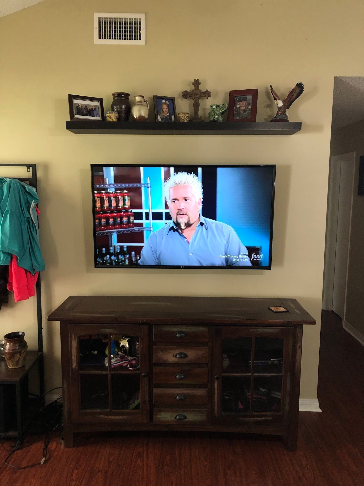 A TV showing Guy Fieri is mounted above a wood cabinet, with a shelf of decor above.