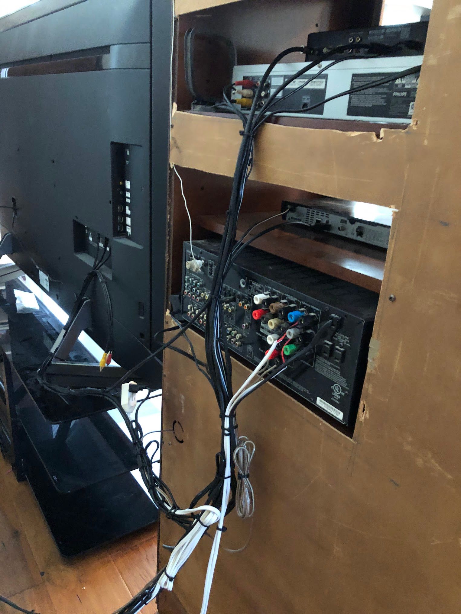 Back of a TV and AV receiver with many connected wires inside a wooden cabinet.