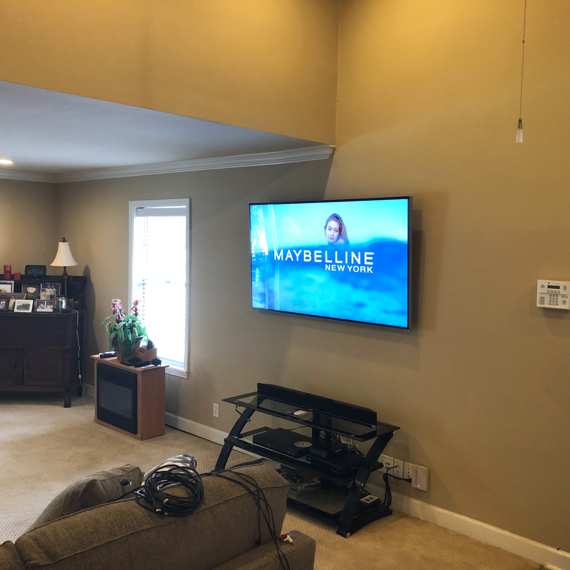 TV showing Maybelline ad mounted on a tan wall, over a black entertainment center.