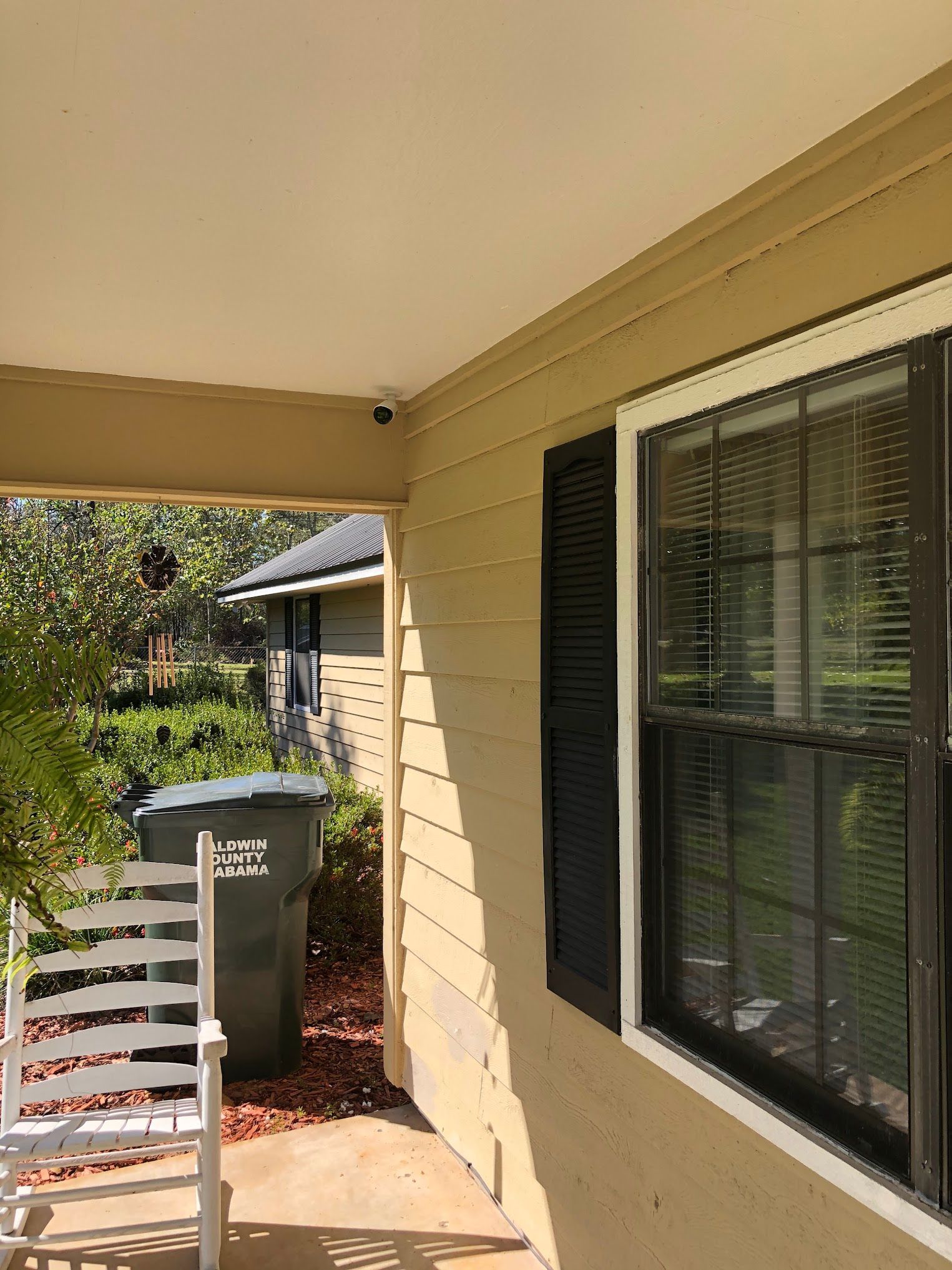 Exterior view of a house porch with white rocking chair, trash can, and black window shutters.