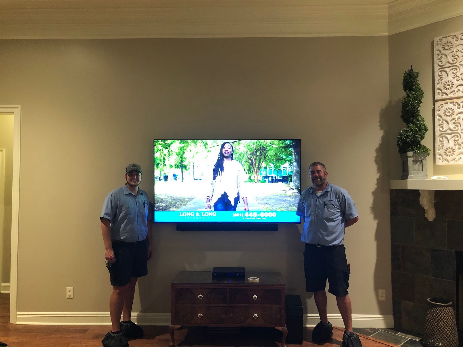 Two men stand beneath a mounted TV showing a woman outdoors, in a living room. Dark wooden cabinet beneath TV.