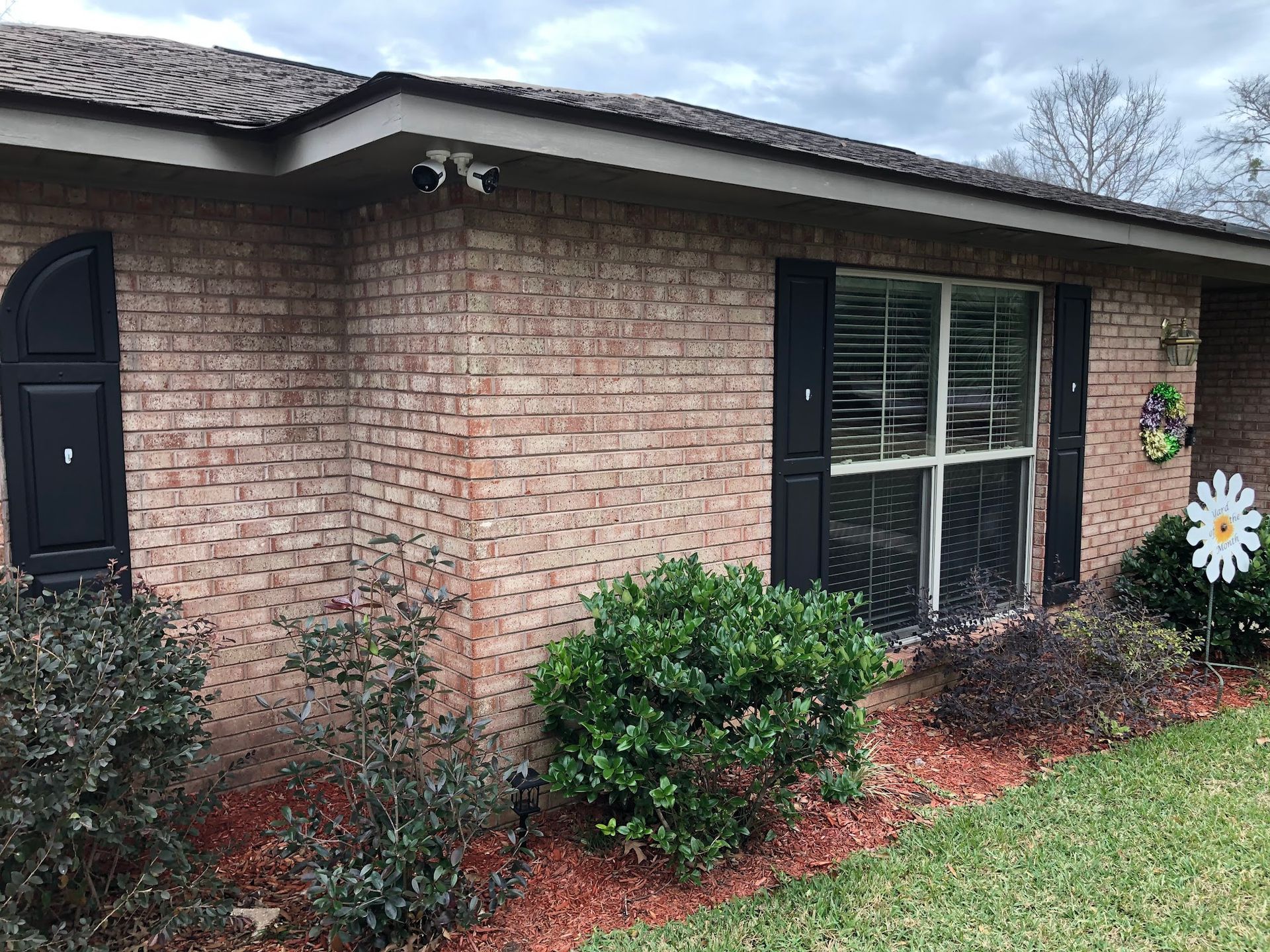 Brick house with black shutters and security cameras, a window, and landscaping with red mulch.