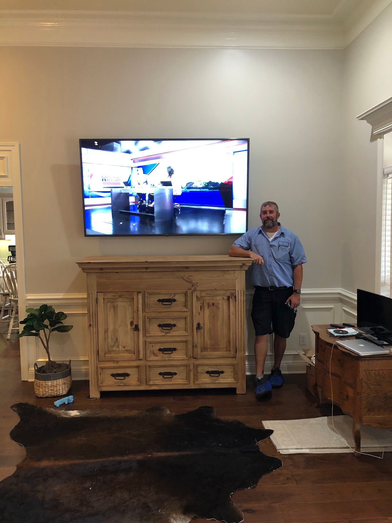 Man stands by a TV mounted above a wooden cabinet in a living room; dark brown rug on the floor.