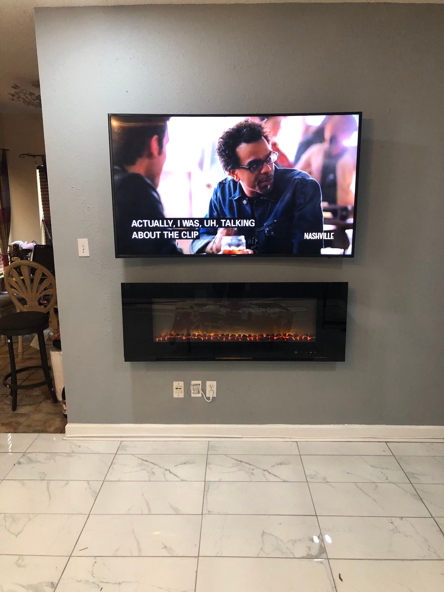 A wall-mounted TV over an electric fireplace, both on a gray wall above white tile flooring.