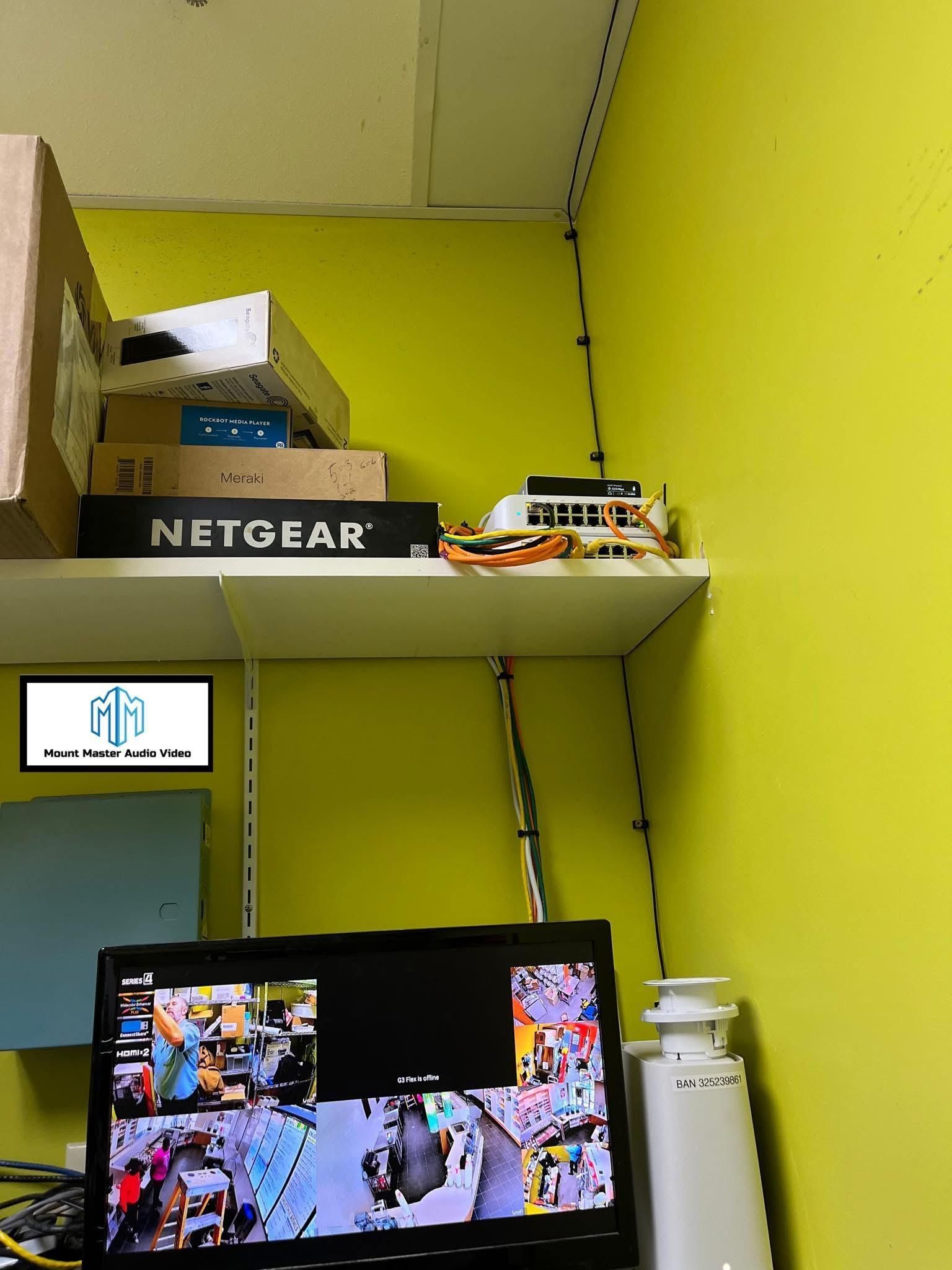 A surveillance monitor showing multiple camera views, below a shelf with routers and equipment.