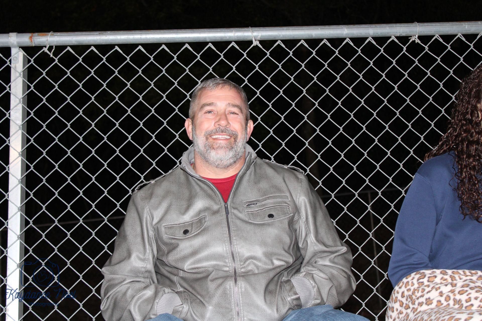 Man with a gray beard smiles, wearing a gray jacket and red shirt, seated near a chain-link fence.