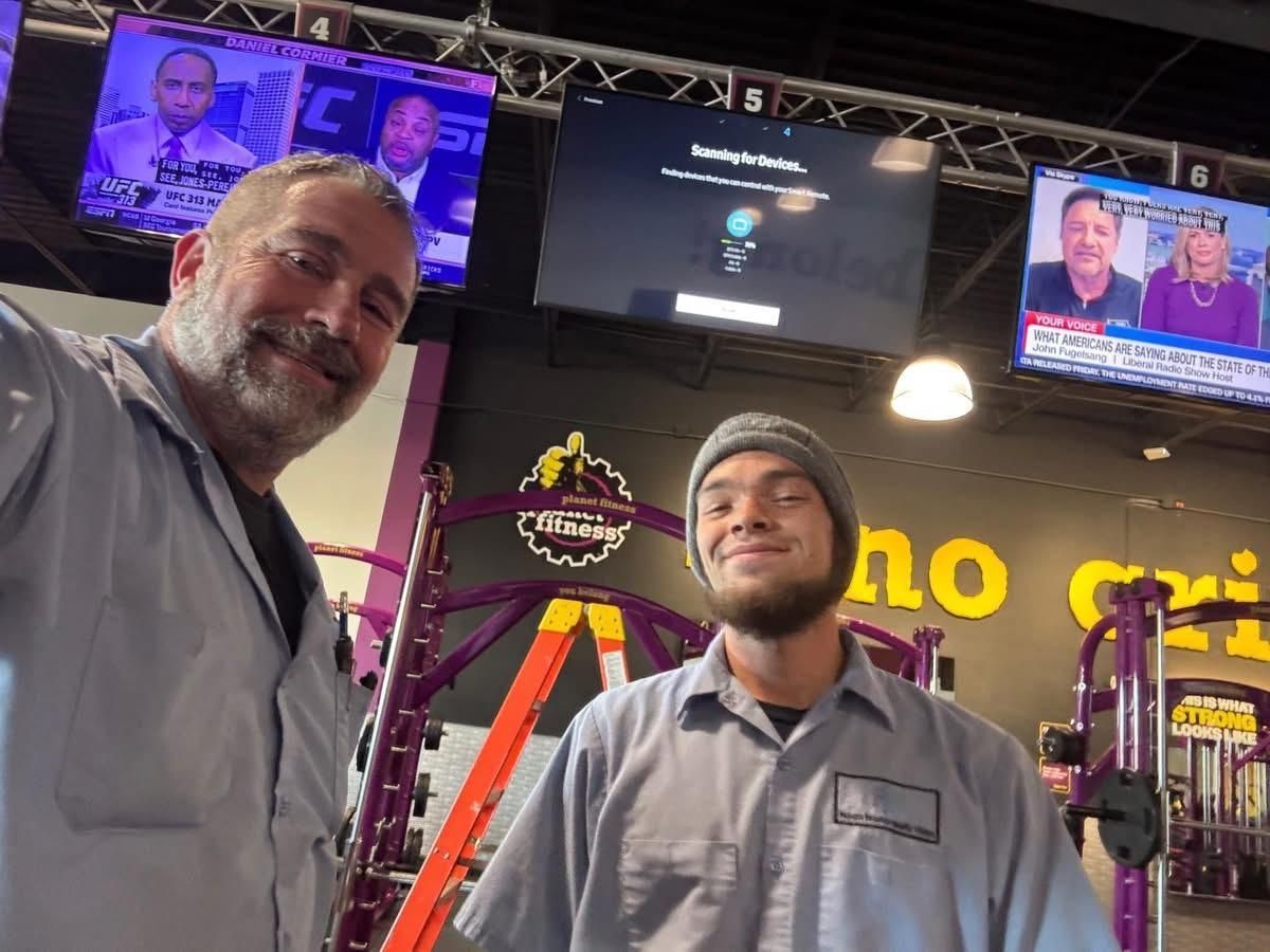 Two men in grey work shirts at Planet Fitness, smiling near equipment and TVs.