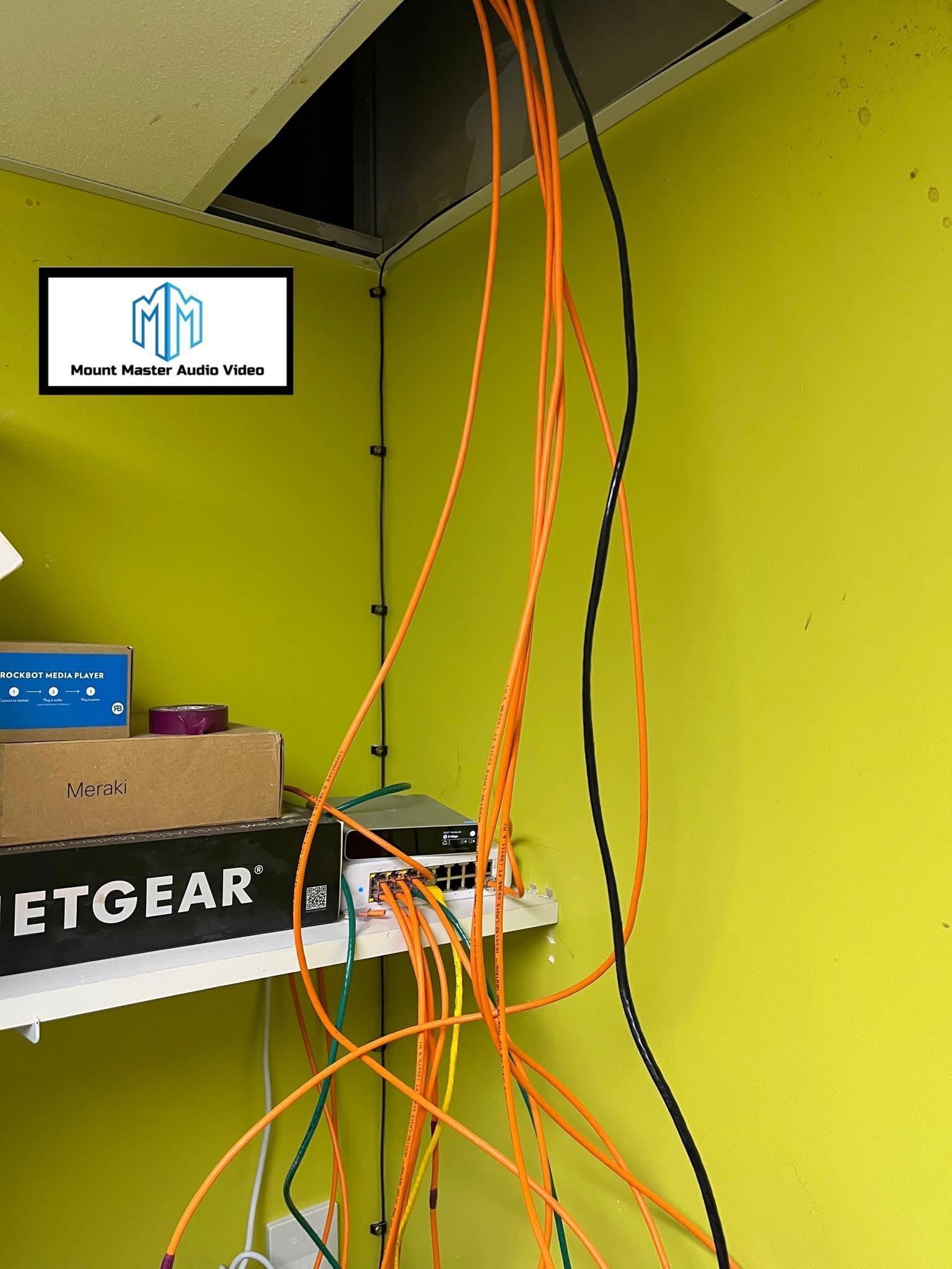 Orange network cables hang from a ceiling access panel, with a Netgear switch on a shelf, against a yellow wall.
