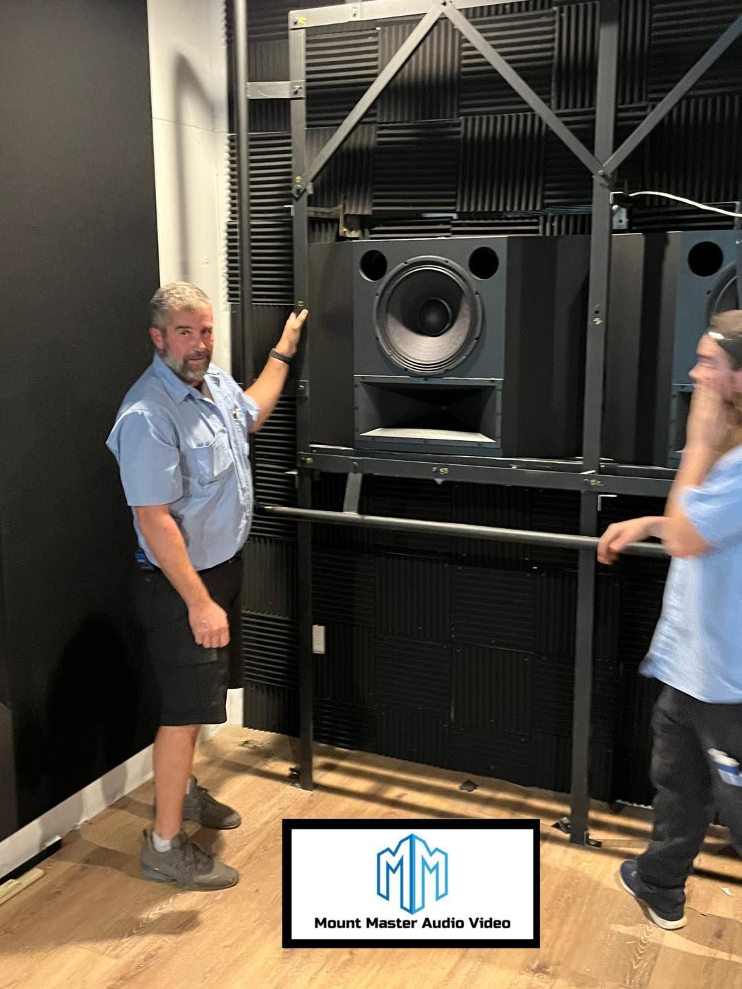 Man pointing to a large speaker in a studio; black frame and walls, other man standing, logo at bottom.