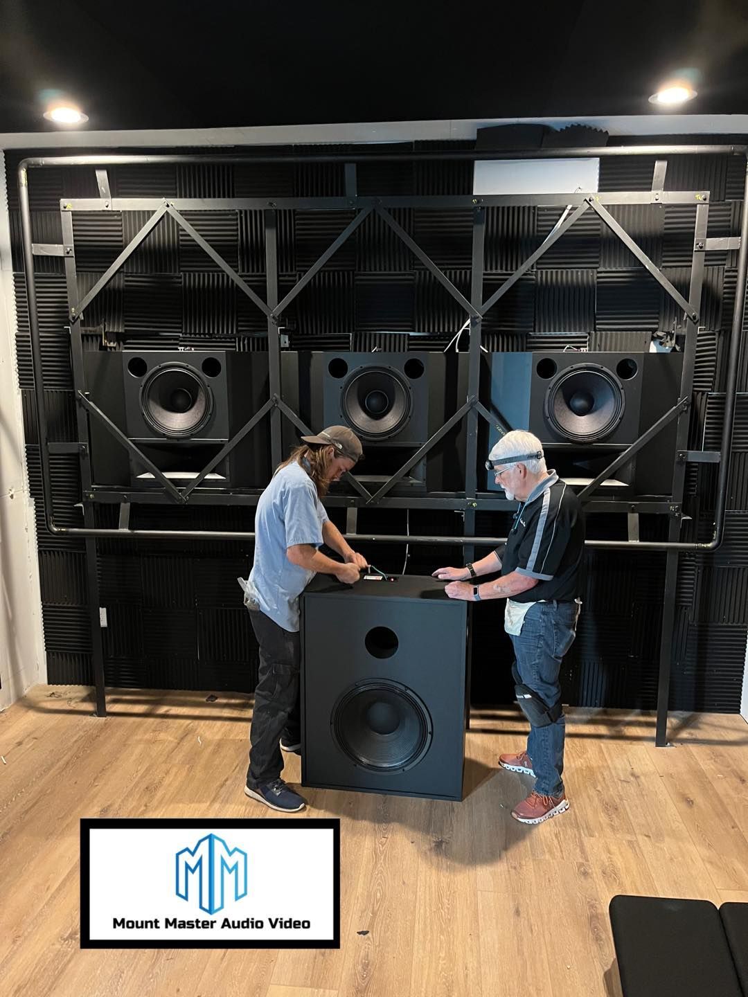 Two men installing a large subwoofer in a soundproof room with other speakers on a metal frame.