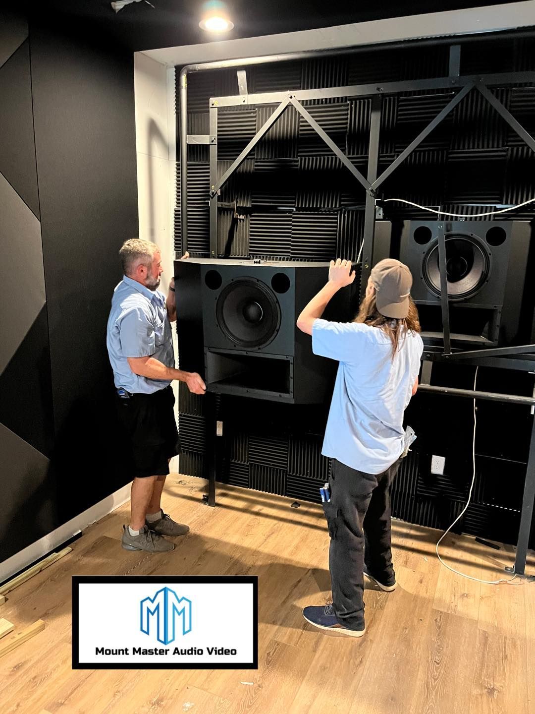 Two men install a large black speaker in a soundproof room.