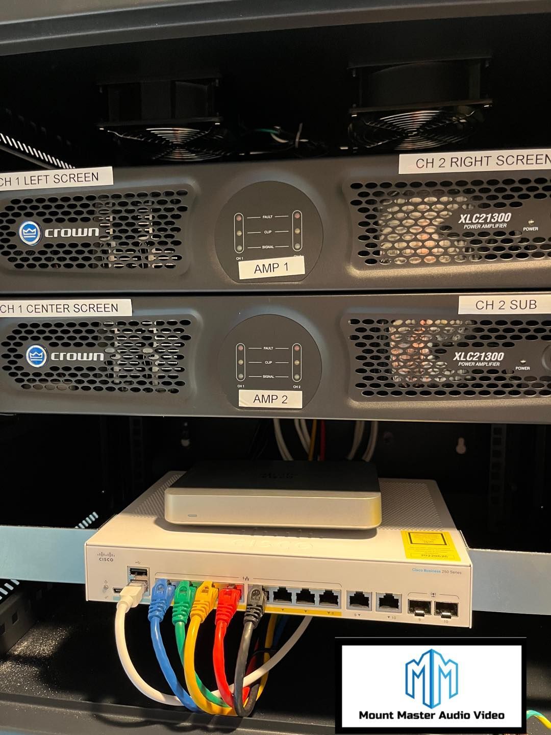 A server rack with audio equipment. Black amps, a white switch, and colored ethernet cables.