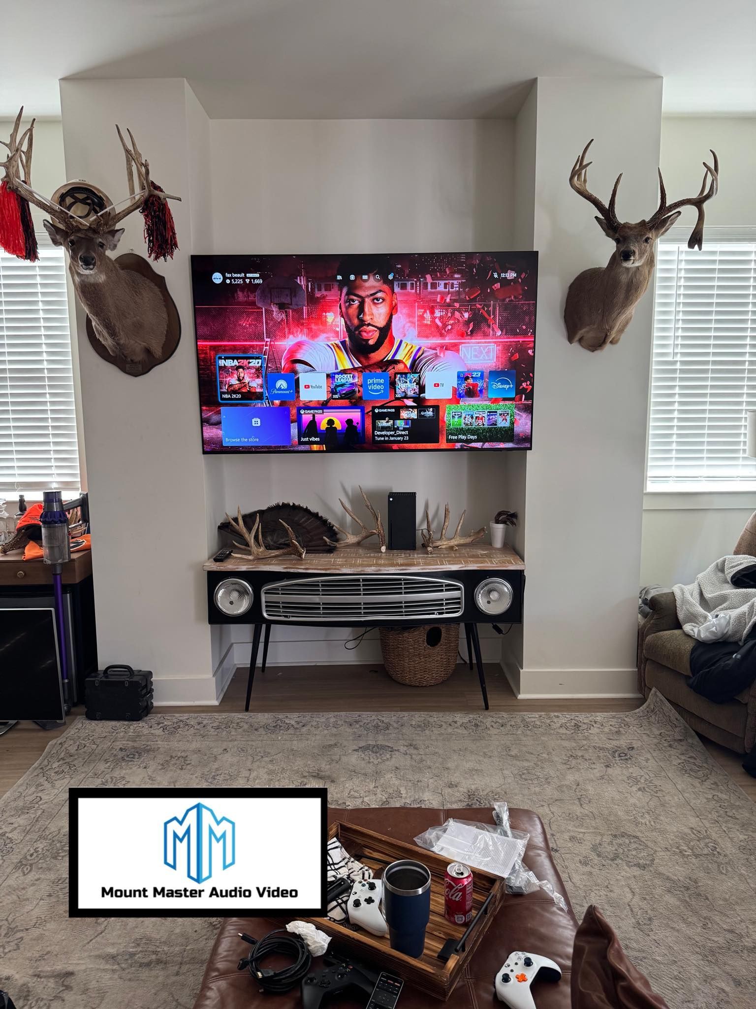 A TV above a cabinet, between two deer heads, in a living room with a rug.