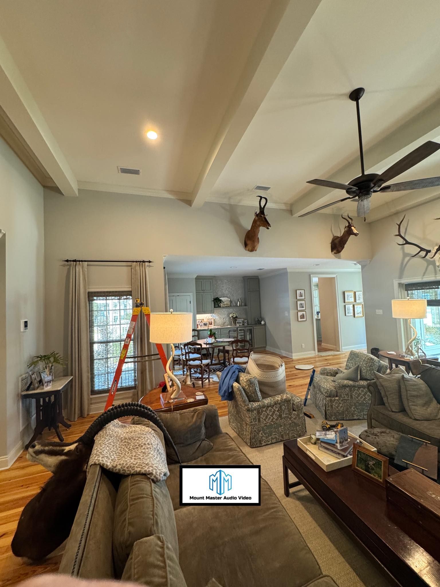 Living room with gray walls, sofas, ceiling beams, deer head mounts, open to kitchen.
