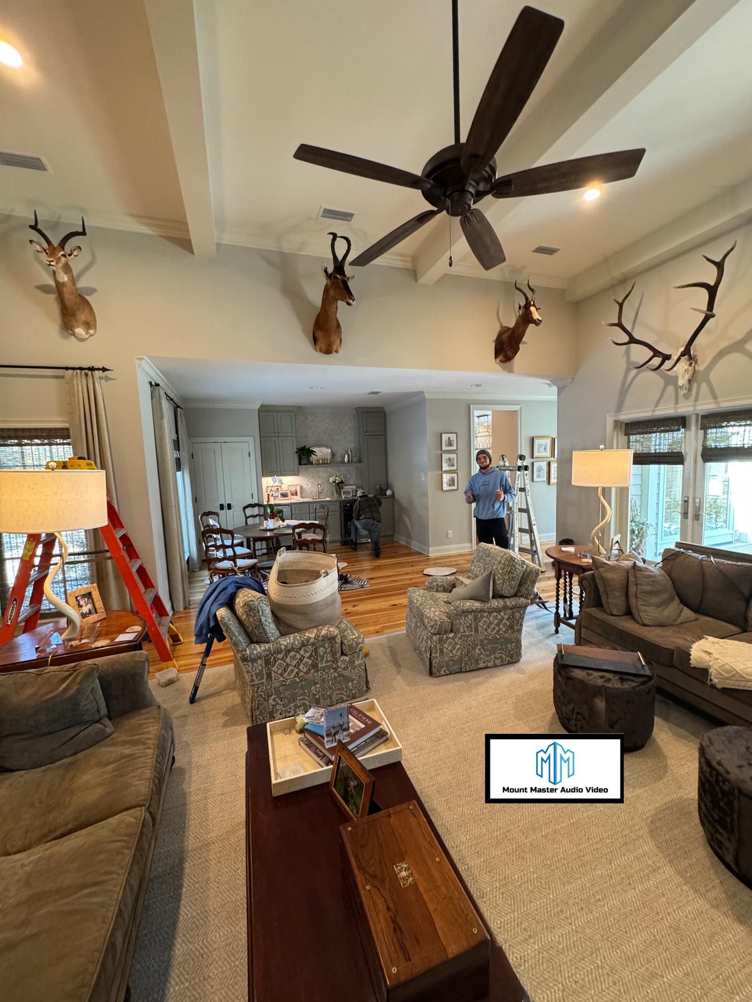 Living room with taxidermied deer heads, ceiling fan, and people working.