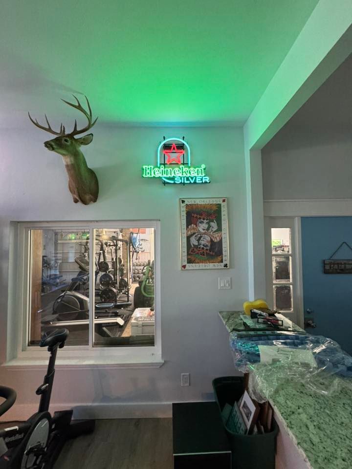 A room with a neon Heineken sign, mounted deer head, and a window showing gym equipment.