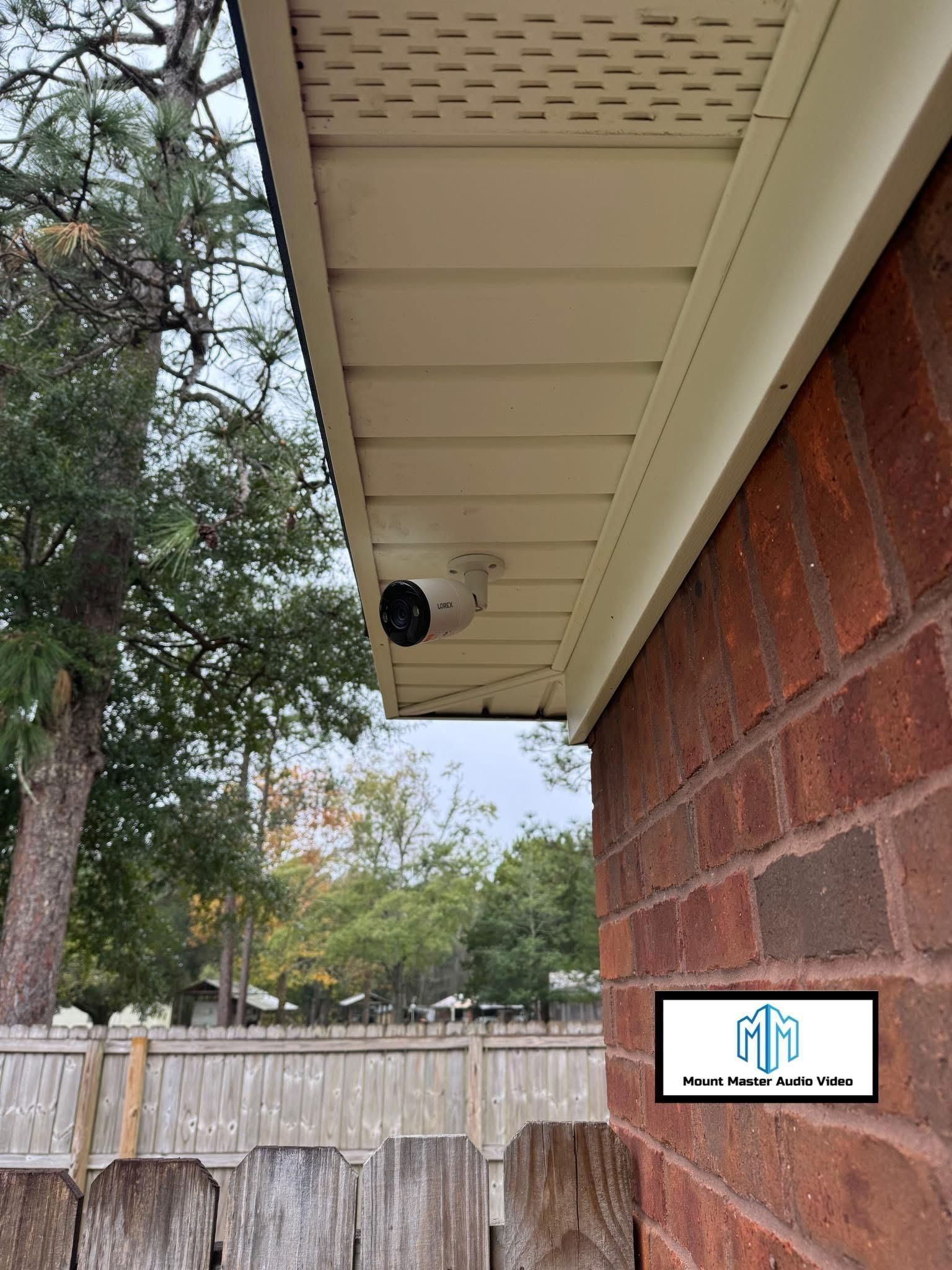 Security camera mounted on beige siding, near a brick wall, overlooking a wooden fence and trees.
